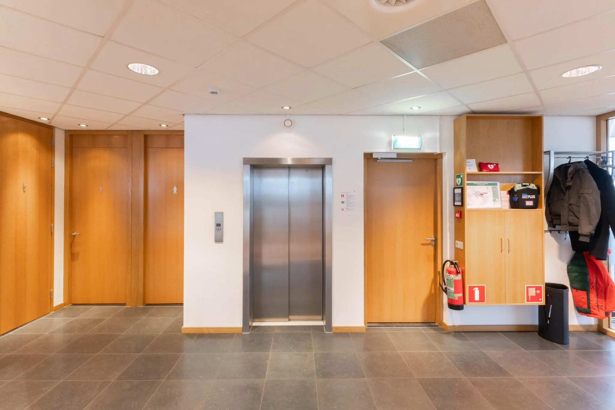 Hal met lift, drie houten deuren, brandblusser en kapstok in een kantoorgebouw aan de 1e Hogeweg.